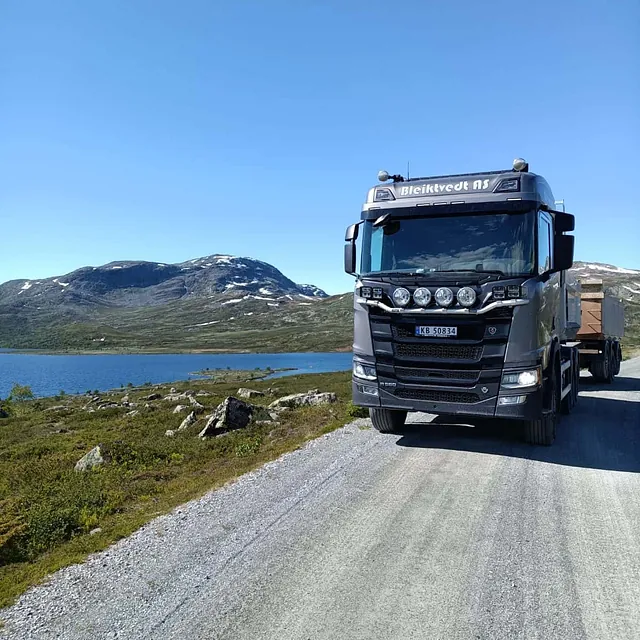 Lastebil fra Bleikvdt AS på grusvei ved innsjø med fjell i bakgrunnen.