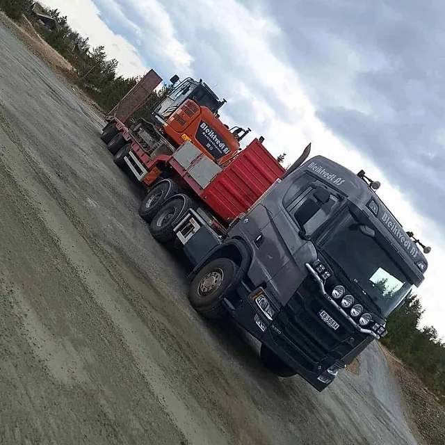 Lastebil frakter oransje gravemaskin på tilhenger over gruslagt plass.