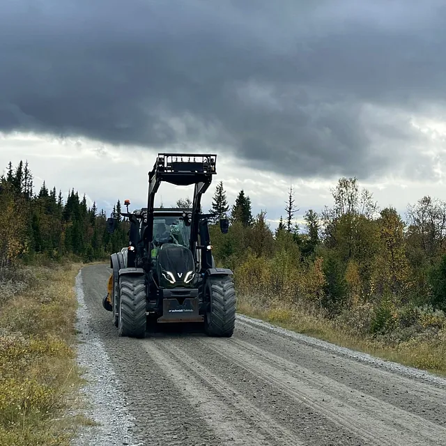 Traktor med frontlaster kjører på grusvei omgitt av høstfargede trær og mørke skyer.