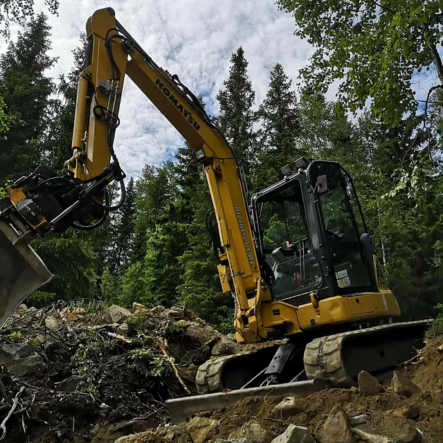 Gul Komatsu gravemaskin jobber i skråning med stein og røtter i skogsterreng.