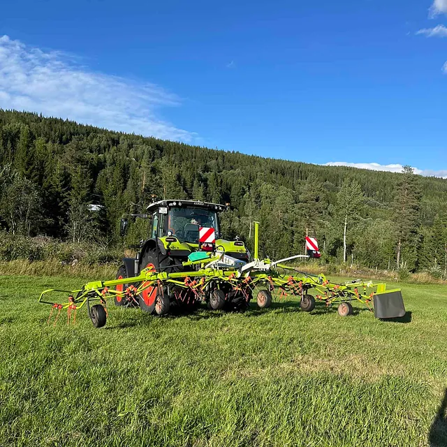 CLAAS-traktor med høyvendere på jorde omgitt av skog og blå himmel.